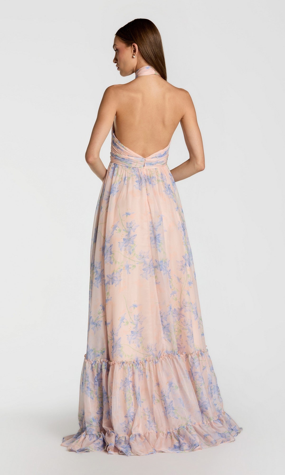 A woman wears the Mac Duggal 68101 long print halter formal dress with a light pink and blue floral pattern, standing and facing away from the camera in an elegant evening look.